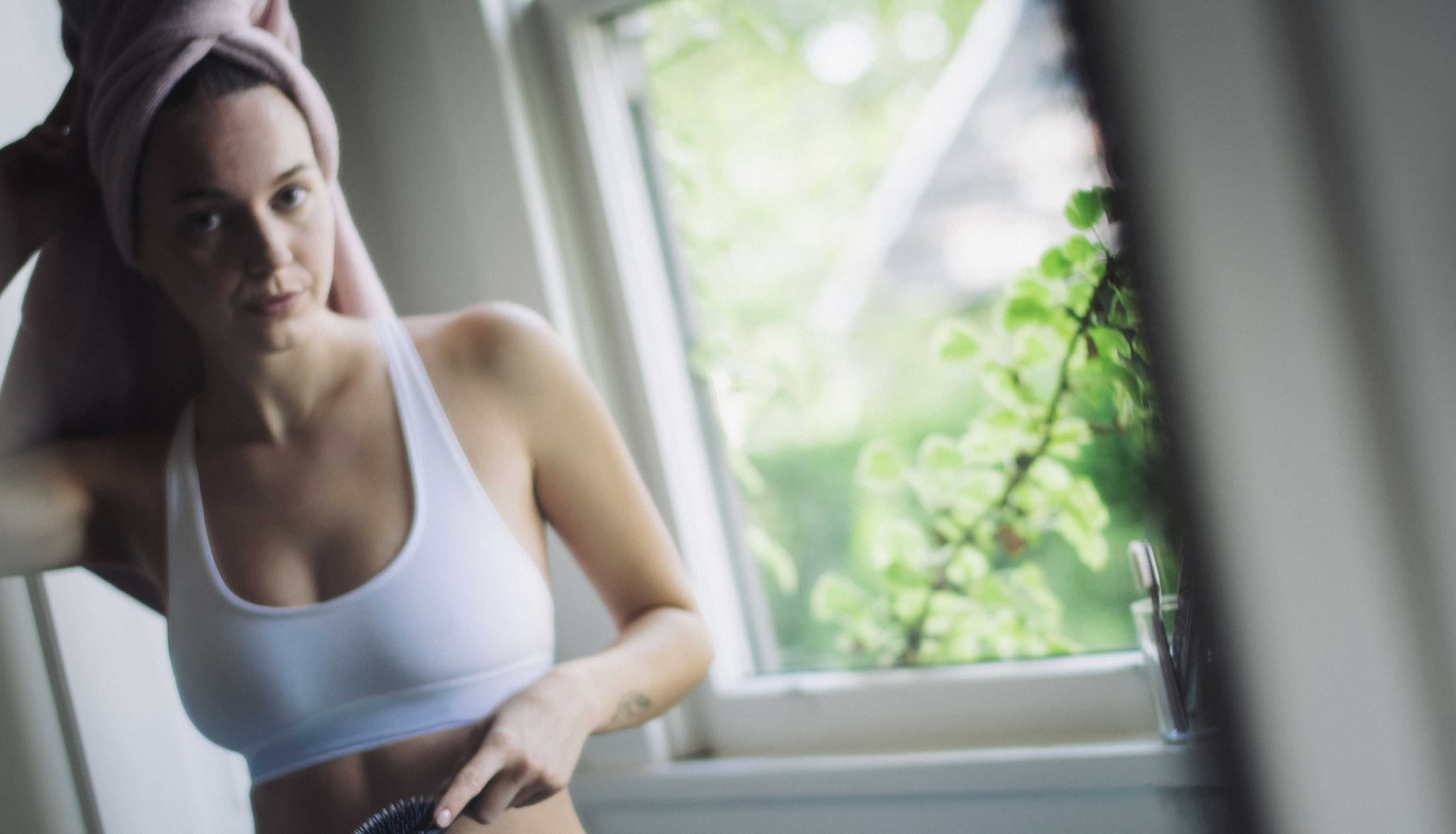 Woman in a white sports bra and leggings standing by a window with greenery outside.