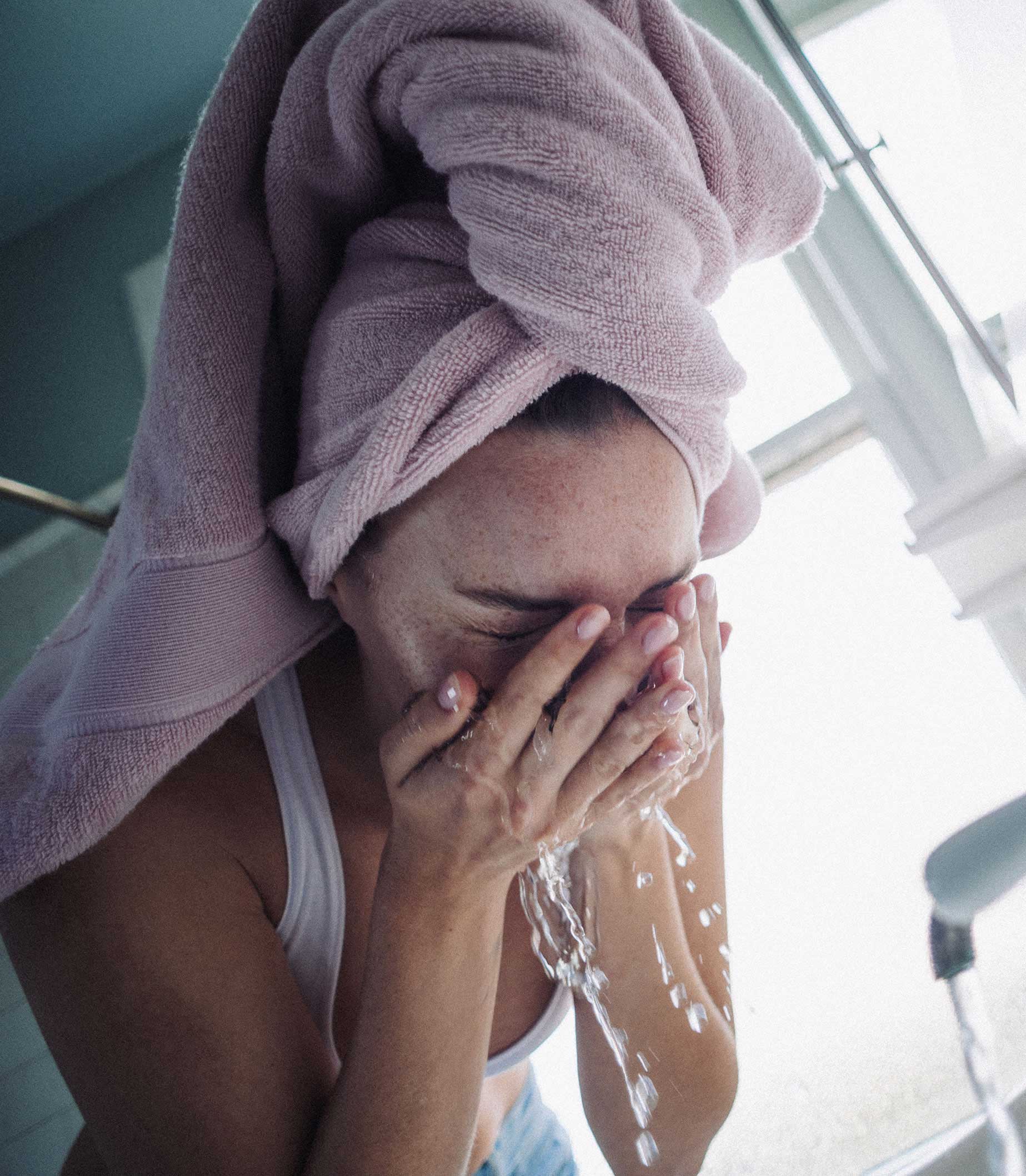 Woman washing her face with water in a bathroom, wearing a pink towel on her head.